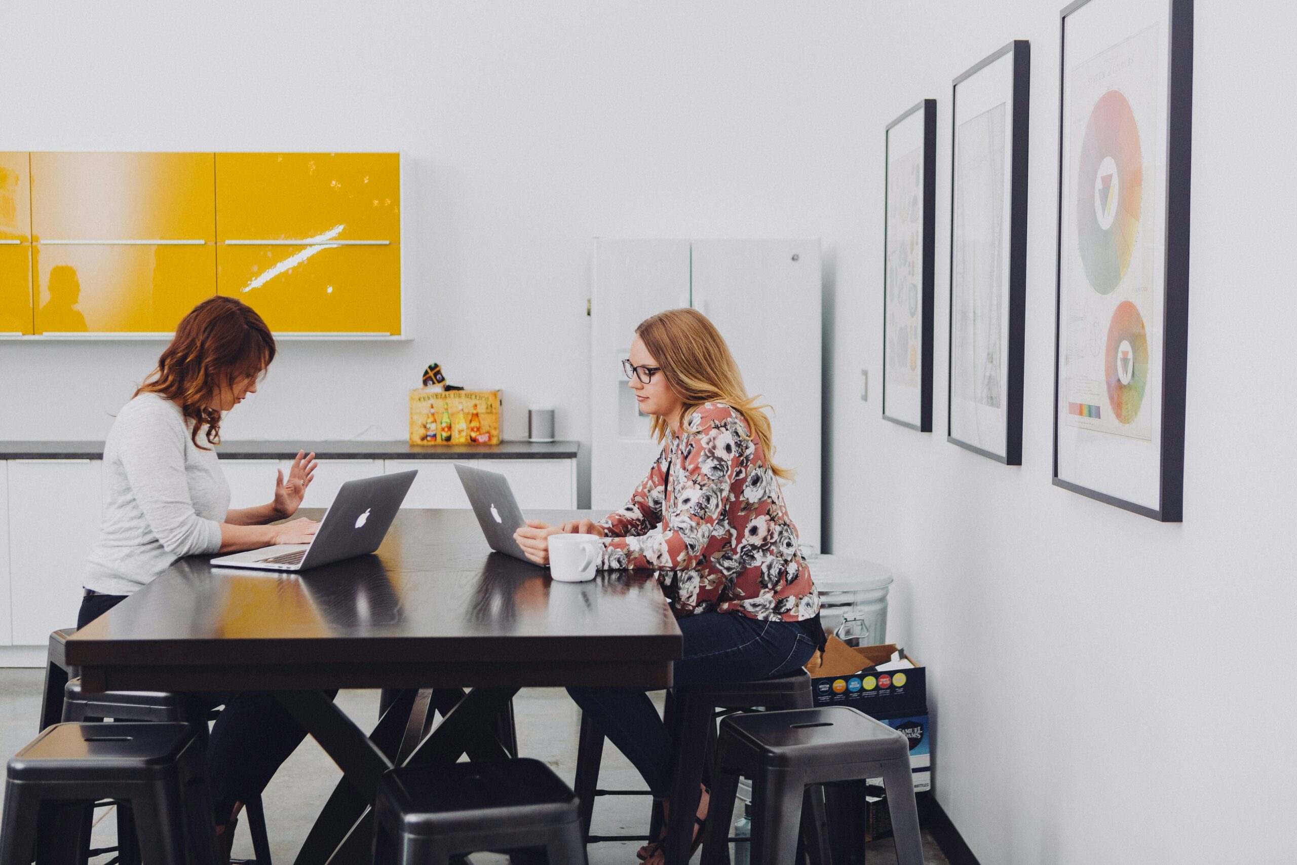 Two women working on laptops at a table with stools in an office